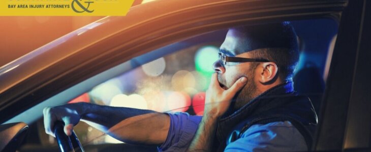 A man yawns while driving a car at night, visibly tired, with blurred city lights in the background.