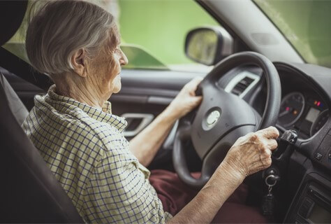 An elderly woman sitting in the driver's seat of a car, holding the steering wheel with both hands.