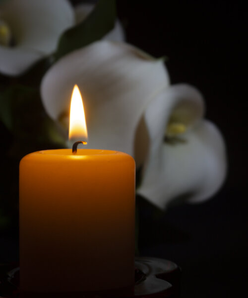 A close up of an orange candle and flame and lily flowers on a dark background.