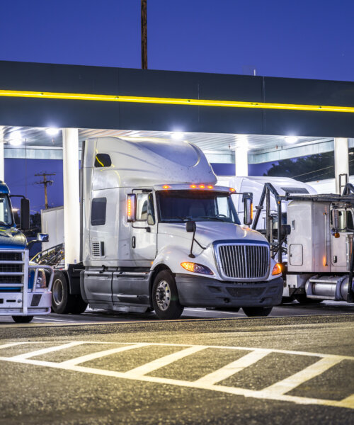 Different make and models big rigs semi trucks with semi trailers standing on the truck stop parking lot under the lighted shelter in night and comply with the movement according to the schedule