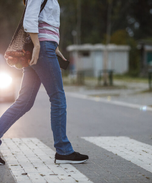 Cut out shot of unrecognizable young woman carrying a reusable mesh bag with groceries and crossing the street on the crosswalk. Car headlights in the background.