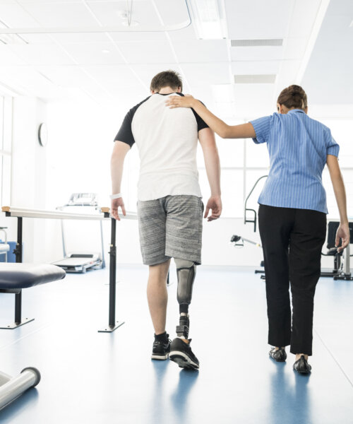 Young man walking with prosthetic limb being assisted by female nurse in hospital. Woman with hand on patient's shoulder.