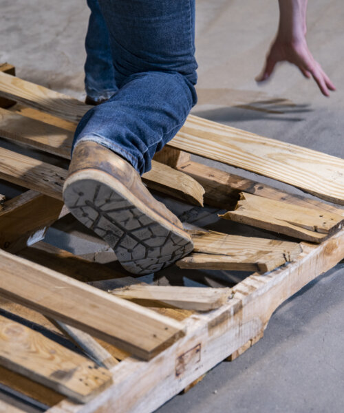 An industrial warehouse, workplace safety topic. A close-up of a person stepping on a broken pallet that poses a severe risk of causing a fall and injury.