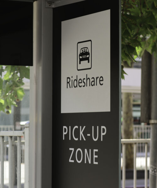 Black and white rideshare pick-up zone sign. Green leaves, metal fence and sign in the background.