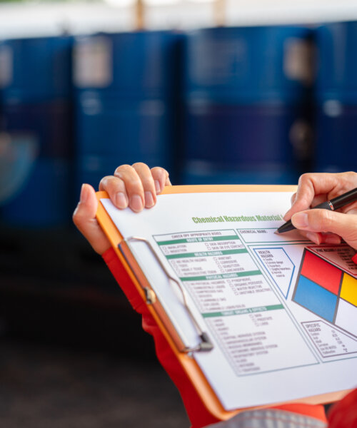 A safety office is checking on the hazardous material checklist form with chemical storage area at the factory as background. Industrial