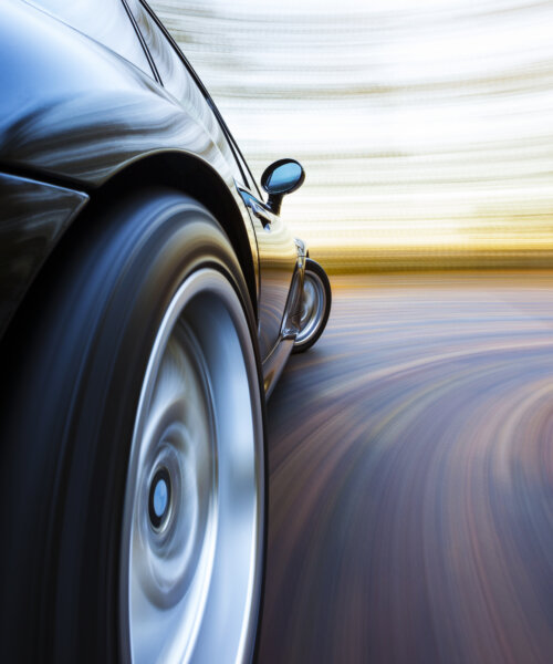 A black sports car turns in to a corner. Autumn leaves are on the road and in the background blur.