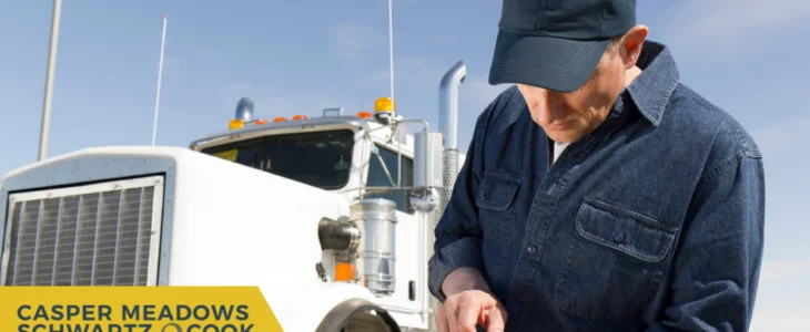 A truck driver in a denim shirt and cap standing in front of a large white semi-truck, focusing on an object in his hands.