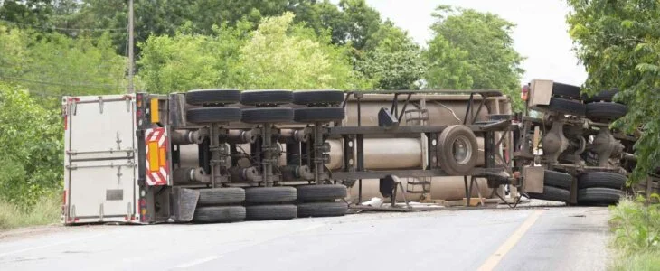 An overturned semi-truck blocking the road after an accident surrounded by trees.