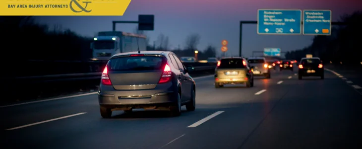 A silver car driving on a highway at dusk, with other cars visible in the background and road signs ahead.