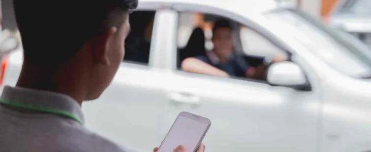 A man holding a smartphone stands near a white car with a driver and passenger inside.