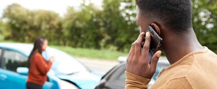 A man on the phone stands near two damaged cars after an accident, while a woman talks in the background.