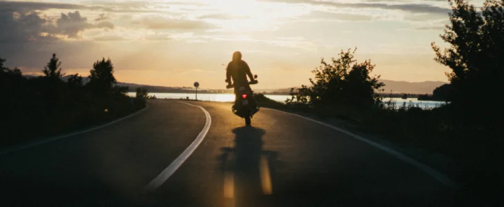 Motorcyclist riding along a curved road at sunset with a lake in the background.
