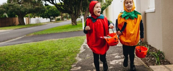 Two young children in Halloween costumes trick-or-treating with pumpkin buckets on a suburban sidewalk.