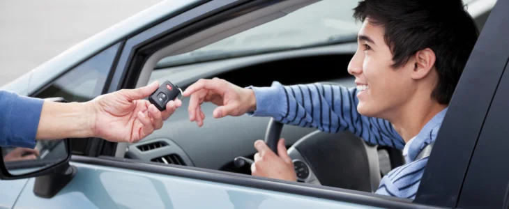 Smiling teenage boy receiving car keys while sitting in the driver’s seat of a car.