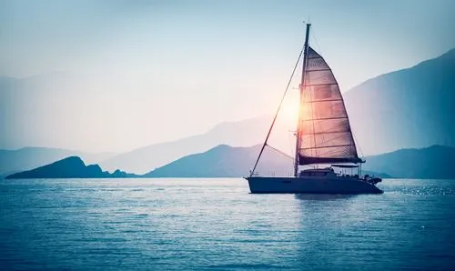 A sailboat gliding on calm water with the sun shining through its sail and mountains in the background.