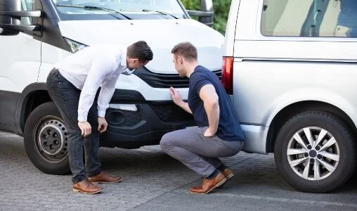 Two men inspecting the front bumper of a white van, possibly discussing a minor collision or damage.