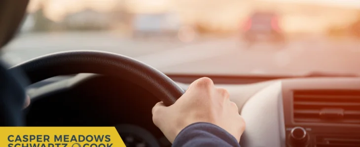 A driver’s hand holding the steering wheel while driving at sunset, with Casper Meadows Schwartz & Cook injury attorneys logo displayed.