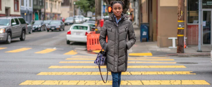 A woman walking across a crosswalk on a city street, wearing a winter coat and holding a handbag.