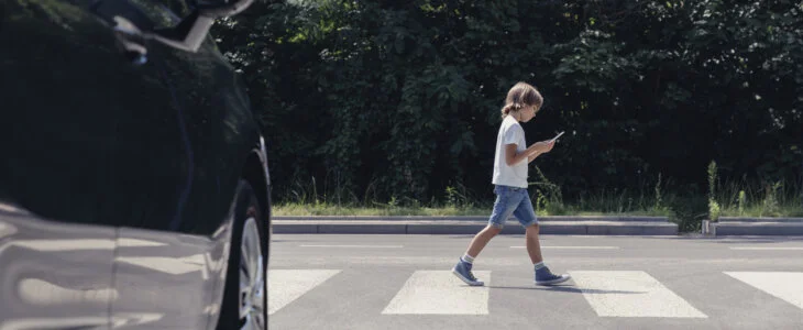 Young boy walking across a crosswalk while looking at his phone, with a car visible on the side of the road.