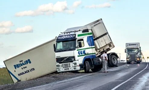 A truck tipped over on the side of the road, with a person standing next to it, while another truck passes by.