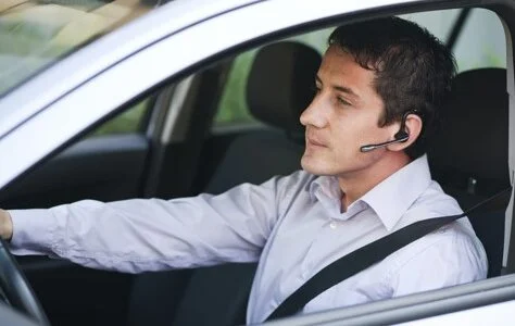 A man in a white shirt driving a car while wearing a Bluetooth headset, with his seatbelt on.