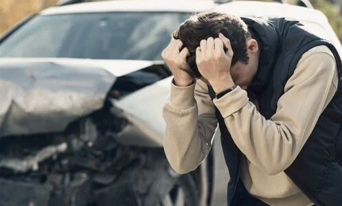 A distressed man holding his head in frustration, standing next to a car involved in an accident with visible damage.