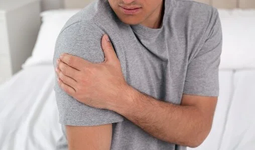 A person with a gray t-shirt holding their shoulder in discomfort, appearing to experience shoulder pain while sitting on a bed.