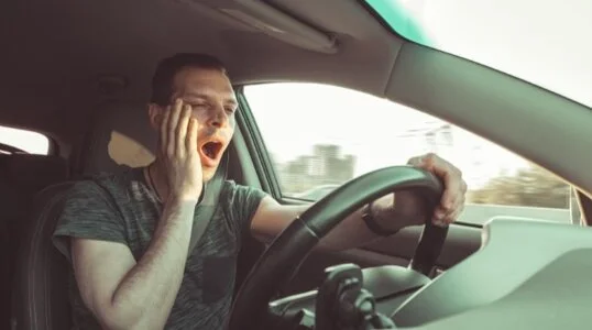 Man yawning while driving, holding his face with one hand as he struggles to stay awake on the road.