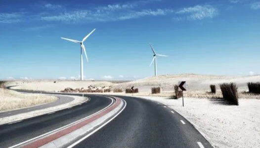 A winding road surrounded by dry, hilly terrain, with two wind turbines standing in the distance under a clear blue sky.