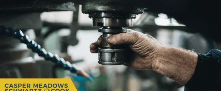 A worker’s hand operating heavy machinery in a factory, with Casper Meadows Schwartz & Cook injury attorneys branding.