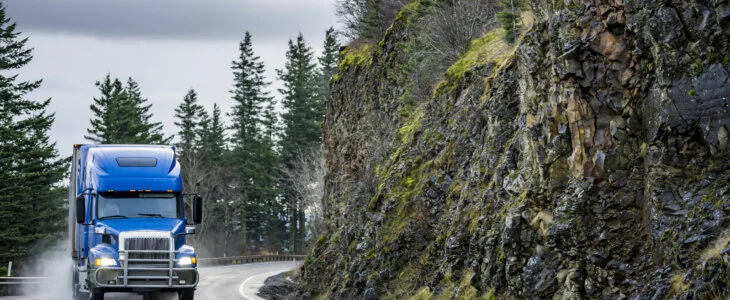 Blue semi-truck driving on a winding mountain road near rocky cliffs
